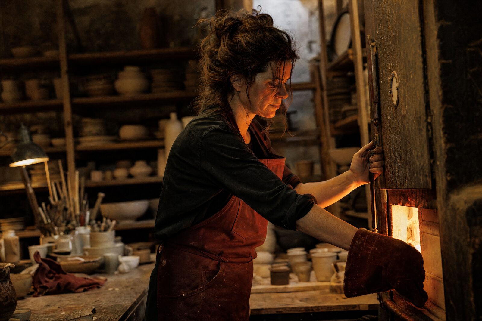 A ceramicist opening a kiln, the glow lighting her face, shelves of bisque-ware behind her