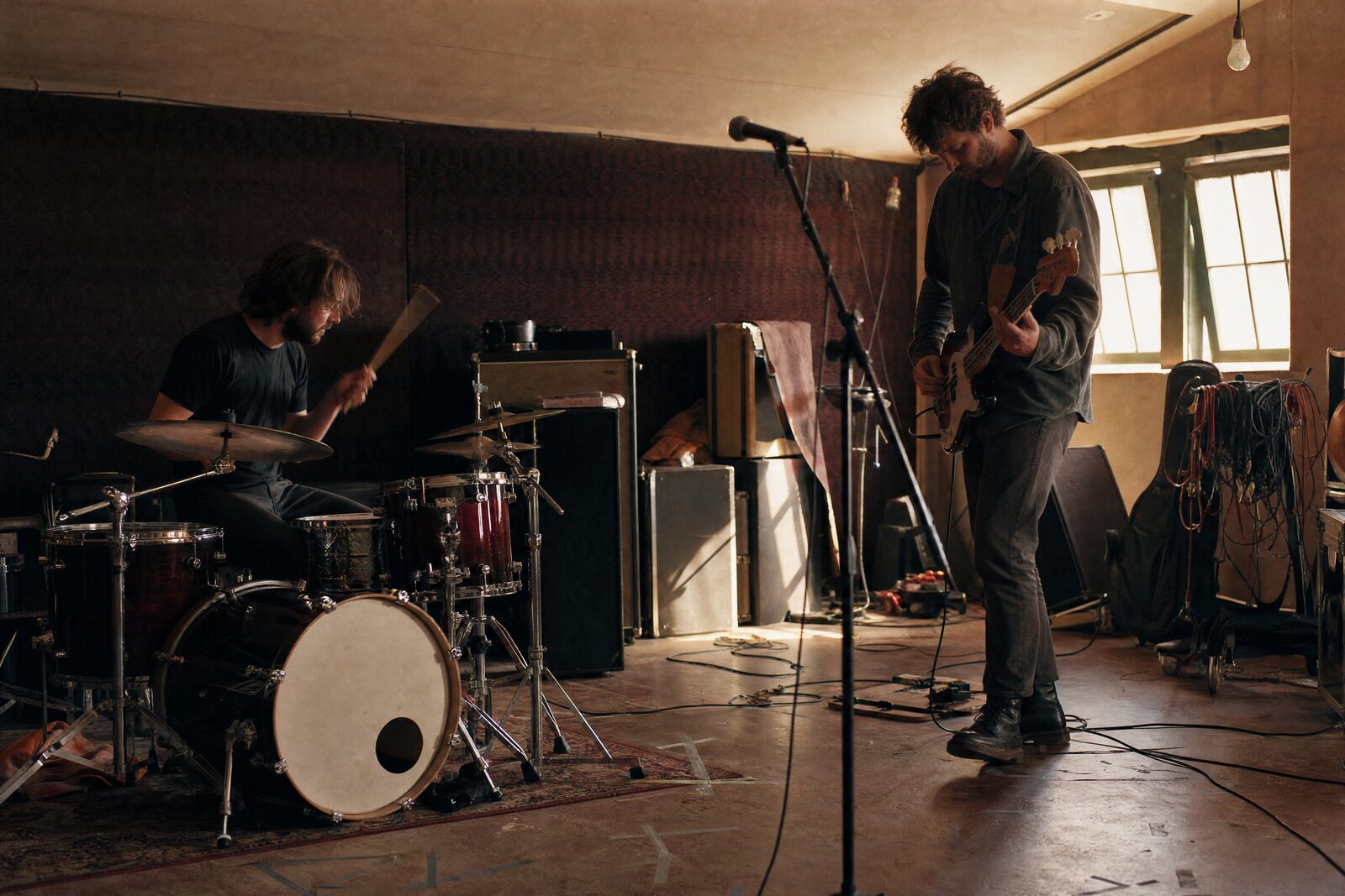A drummer and a bassist mid-take in a rehearsal room, tape marks on the floor and amps stacked, a third mic stand standing empty