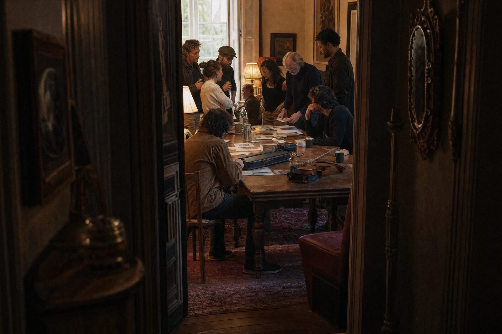 A doorway seen from a dim hall, opening into a warm room where people talk over a long table covered in prints and instruments