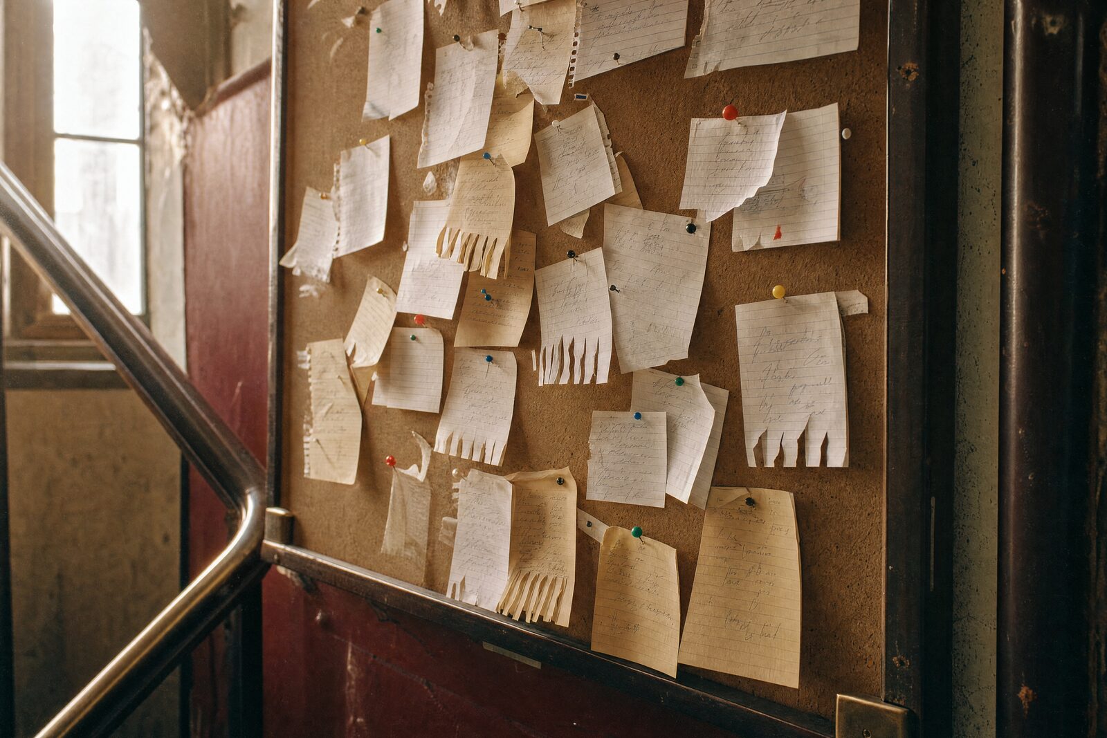 A cork noticeboard in a studio stairwell, thick with handwritten cards asking for a guitarist, a voice, kiln space
