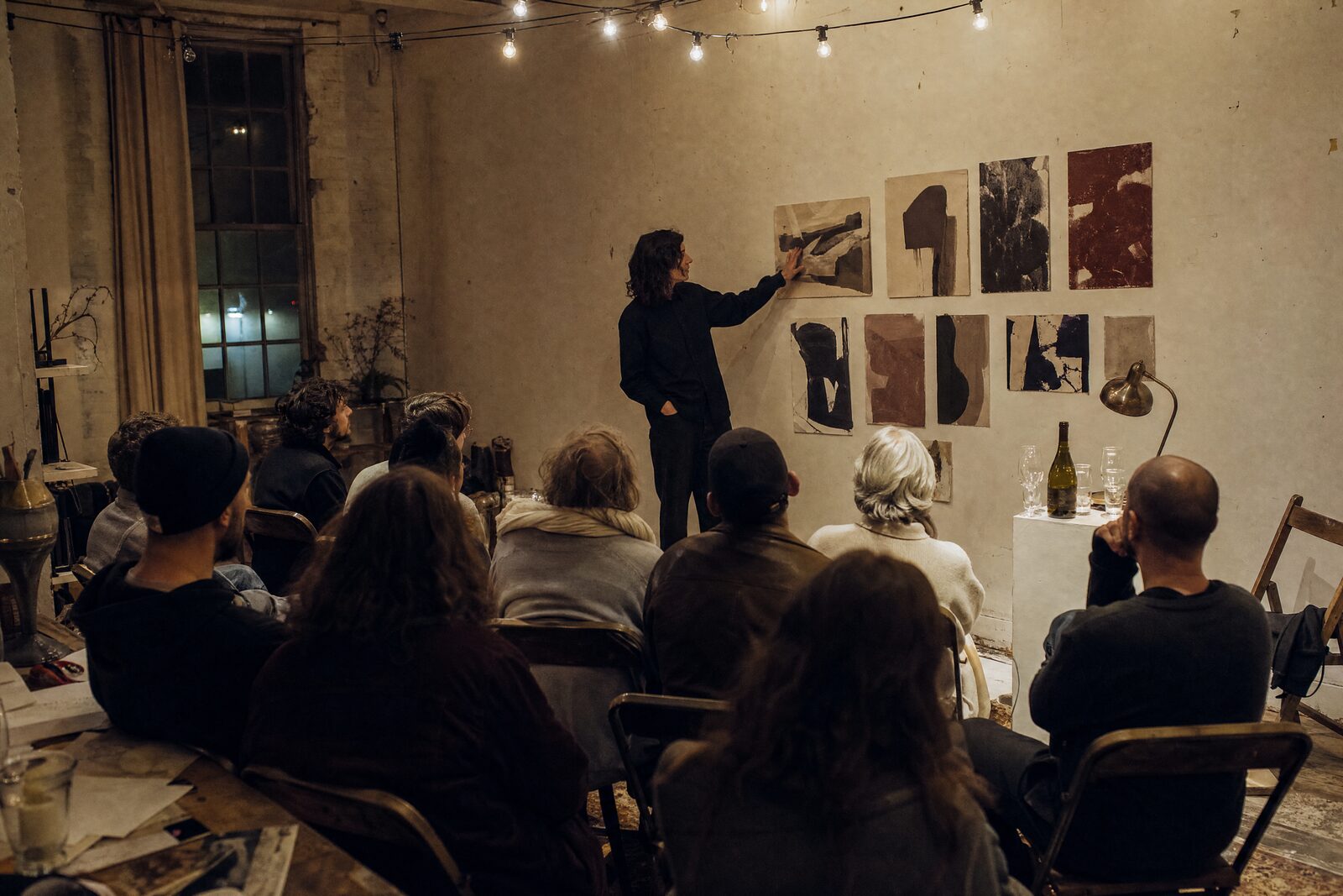 A studio night — someone showing work on a wall to a small crowd, a bottle on a plinth, folding chairs
