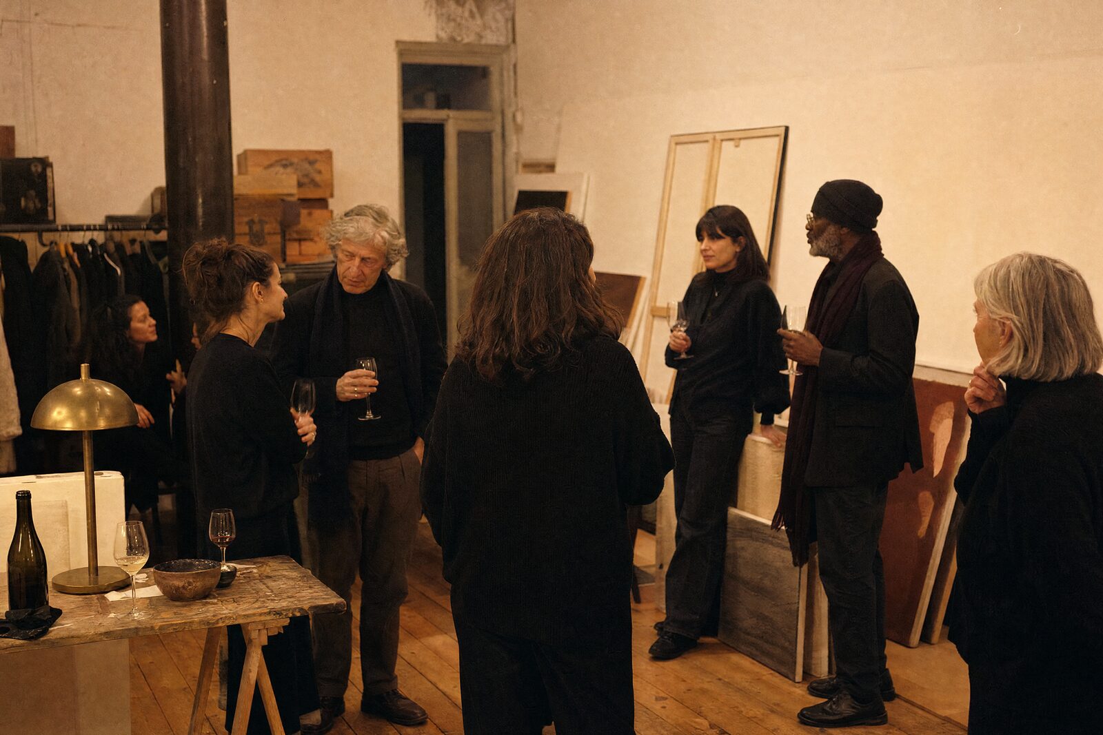 A gallery back room after an opening, a few people in conversation among leaning canvases, glasses half full